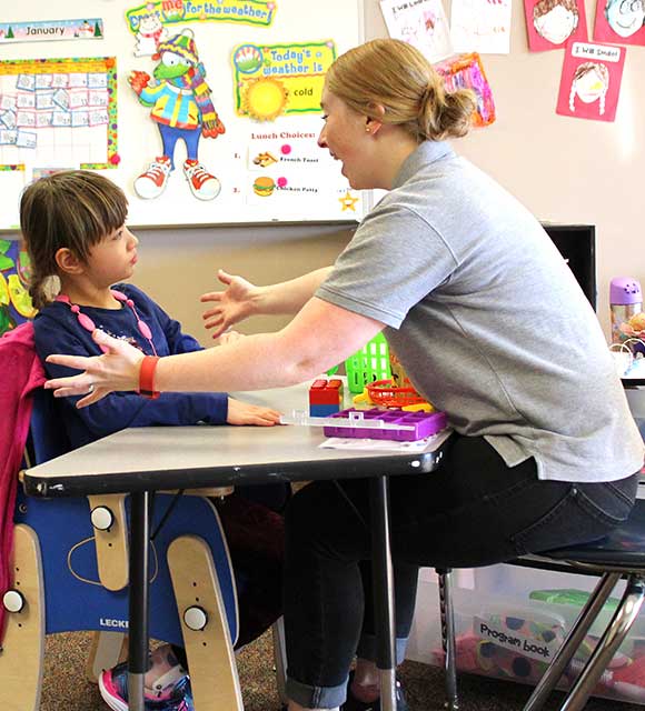 A special education teacher reaches for a hug from a young girl in an autism support classroom.