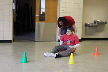 A special education student in a red shirt smiles as his teacher pushes him through an obstacle course