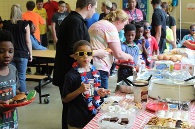 A young boy wears sunglasses and a flowered necklace while others gather food during an event at a special needs school