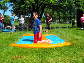 A young boy in a blue shirt looks at the camera from the center of a sprinkler pad
