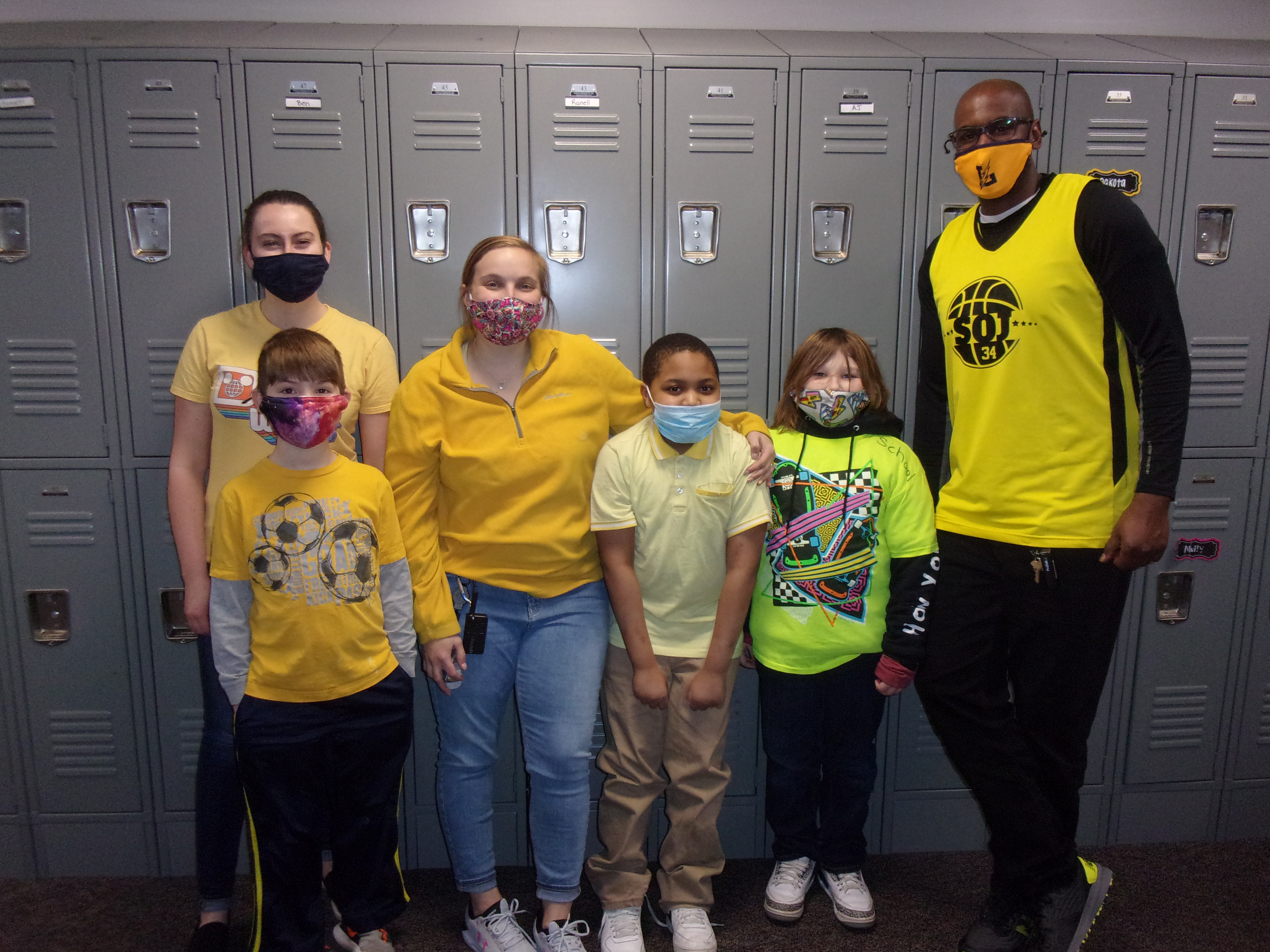 Students and Teachers dressed in shades of yellow pose by a bank of lockers. They're all wearing facemasks