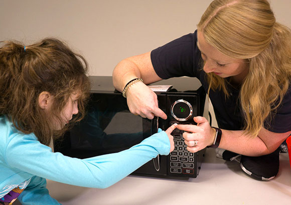 Special education teacher helping elementary school girl with autism develop life skills with a cooking lesson using a microwave to cook popcorn.