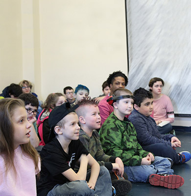 A group of young special education students sits together on the floor while listening to a presentation.