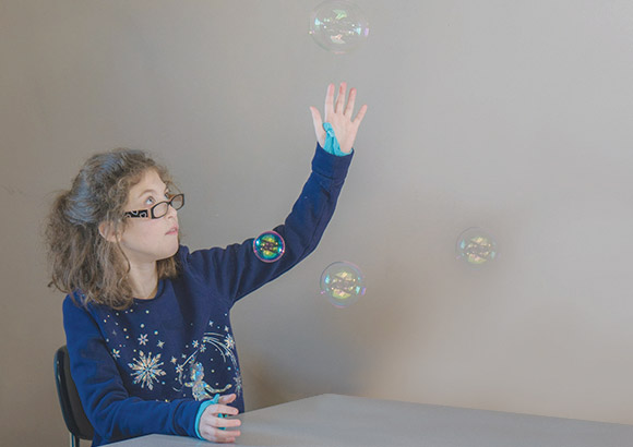 Special education elementary school girl, with autism, practicing counting by popping bubbles blown by staff member.