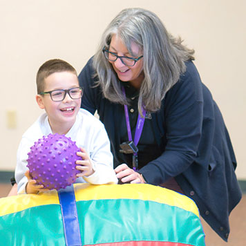 Staff member helping happy special education boy practice his balance by rolling on top of the rocking barrel to retrieve a ball in an occupational therapy session.