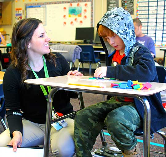 A special education teacher helps her elementary school student with an exercise using colored foam shapes.