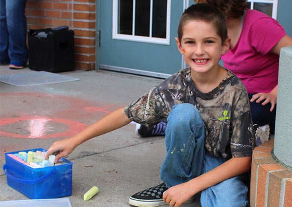 An elementary school boy smiles while drawing with chalk outside his special education school.