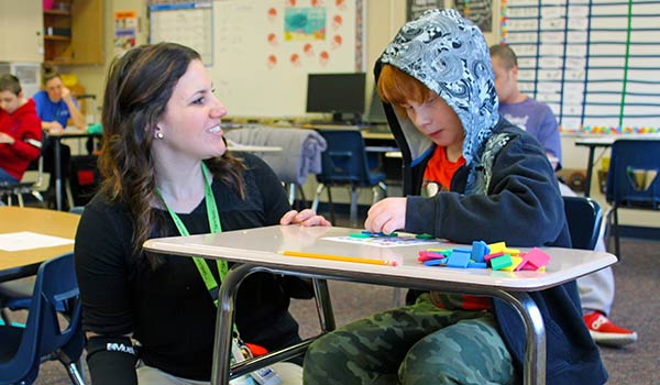 A special education teacher helps her elementary school student with an exercise using colored foam shapes.