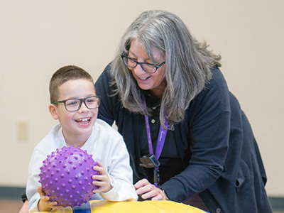 Staff member helping happy special education boy practice his balance by rolling on top of the rocking barrel to retrieve a ball in an occupational therapy session.