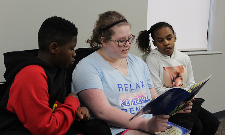 two young students follow along as an older student reads "Black Beauty" to them. 