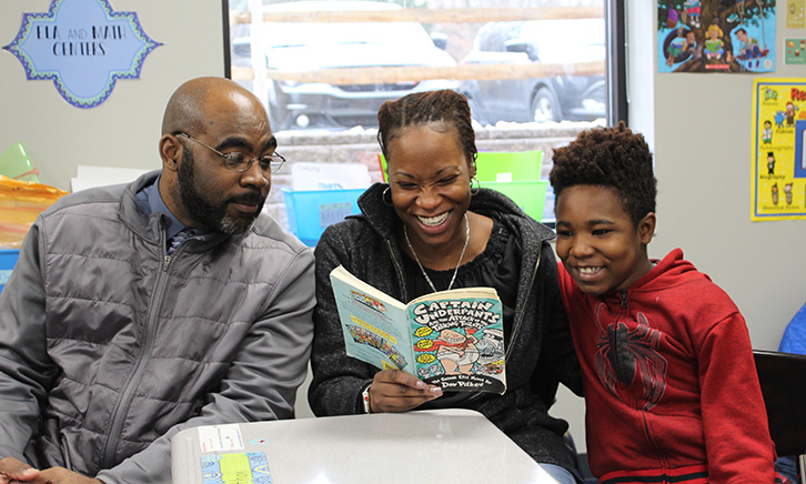 Mother and father read a grade level book called, "Captain Underpants" with their son. The mother is laughing.