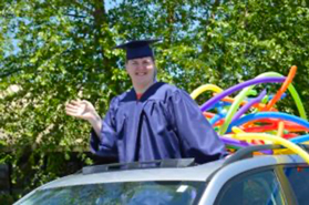Male student in blue cap and gown waves from the sun roof of his family's car. He's grinning ear to ear!