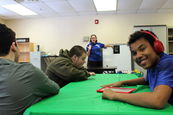 A high school boy in headphones smiles for the camera in his autism support classroom