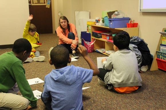 Elementary students sitting in a circle in their special education classroom raise their hands to answer a teacher's question.