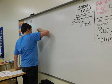 A high school student in a blue shirt writes on the white board of his special needs classroom