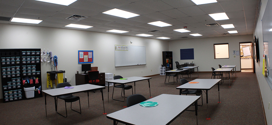 High School Classroom. Wide, plain desks, shelf with tupperwares for sorting projects, white board, well lit, cheerfull. 