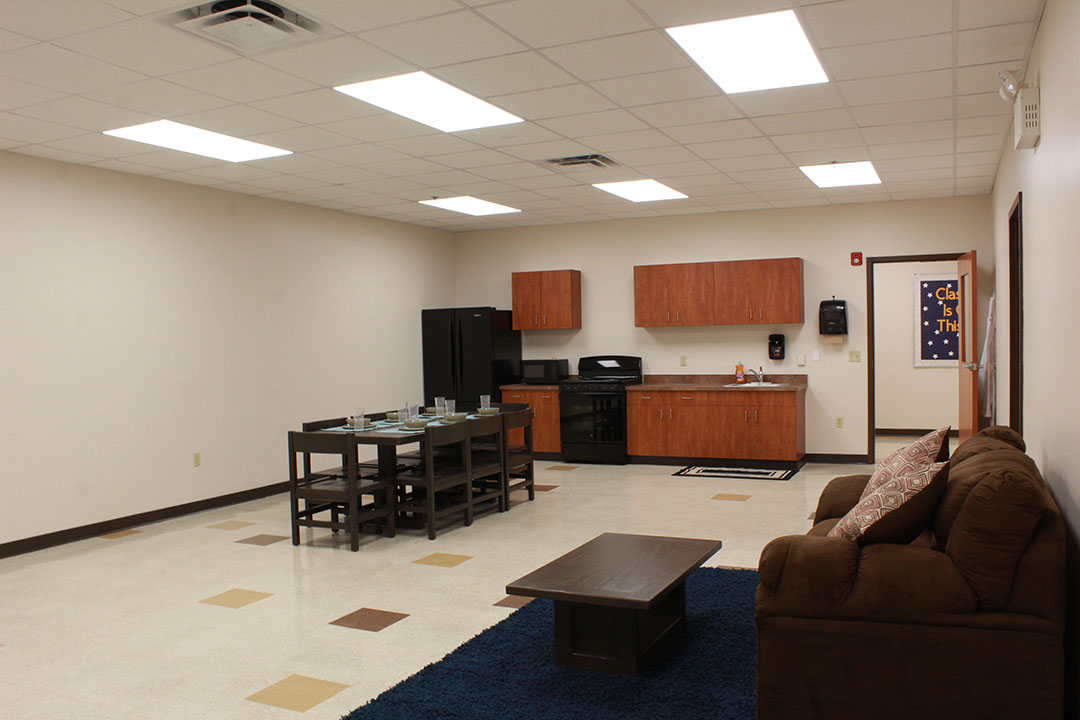 Life Skills Apartment. Brown couch, table and rug, dining table with six places, small kitchen along one wall with stove, cabinets, sink and refrigerator. Not shown: Bed with sheets and nightstand.