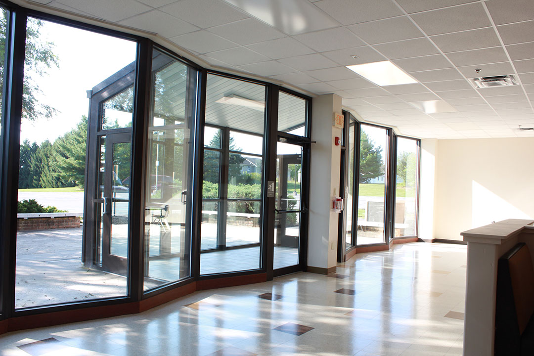 Student Entrance to the School. Floor to Ceiling windows along entire wall leading to glassed in atrium. Sunlight on the floor, spotted shade from trees outside.