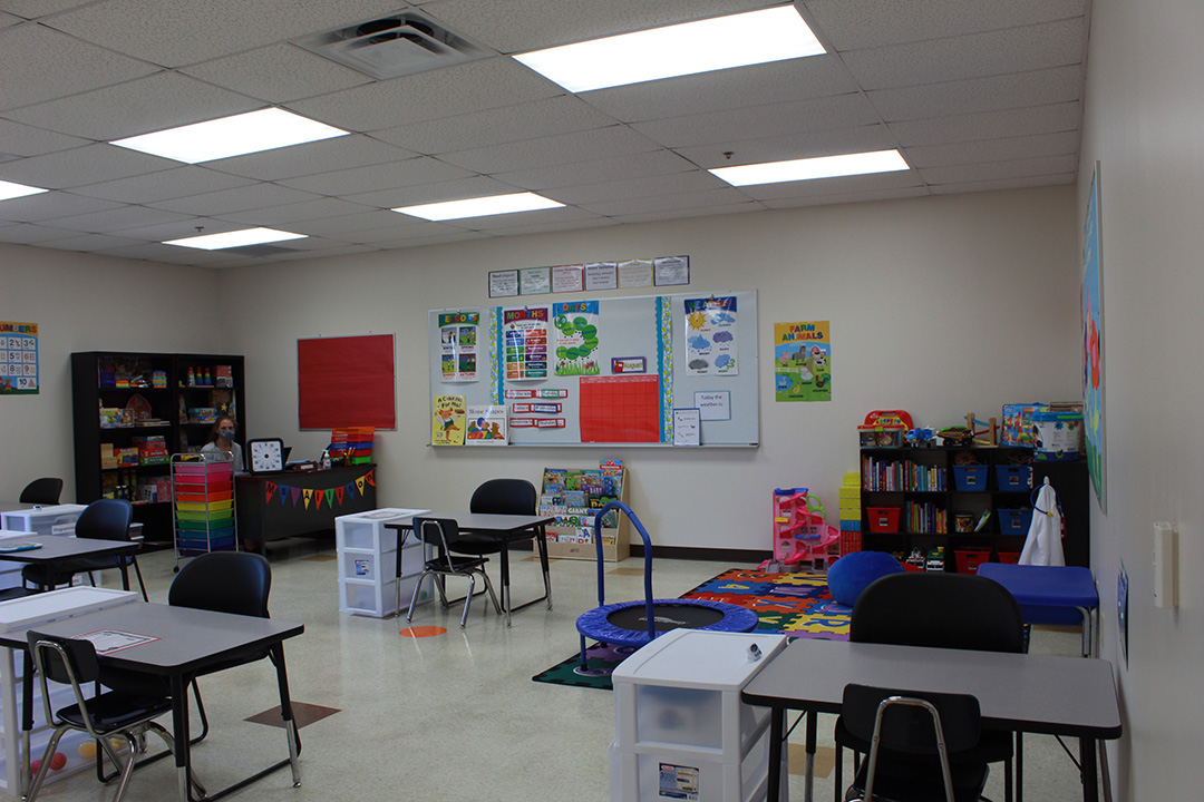 Elementary Age Classroom. Small desks, colorful posters, shelves with bright books. Each desk has a little set of drawers for organizing student materials.