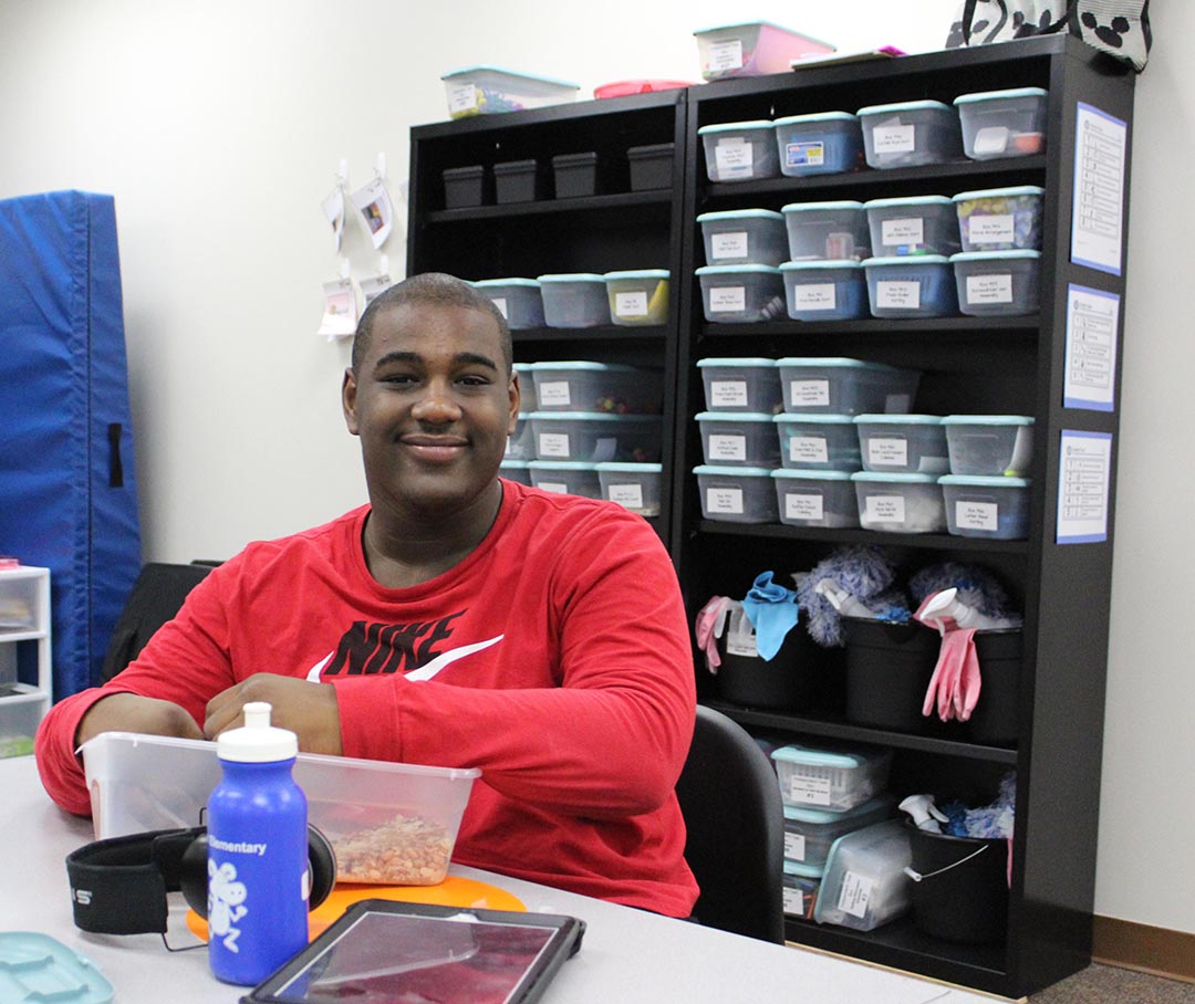 High School Special Education Student in front of shelf full of tubs of materials. Student is holding something in a tub, mid lesson