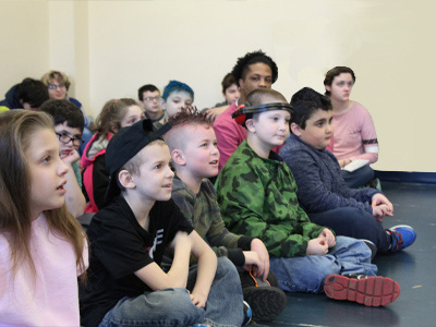 A group of young special education students sits together on the floor while listening to a presentation.