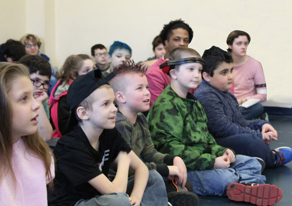 A group of young special education students sits together on the floor while listening to a presentation.