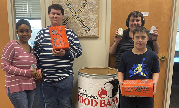 Four Students with cans and non-perishable food items around a big tub that says "Food Bank"