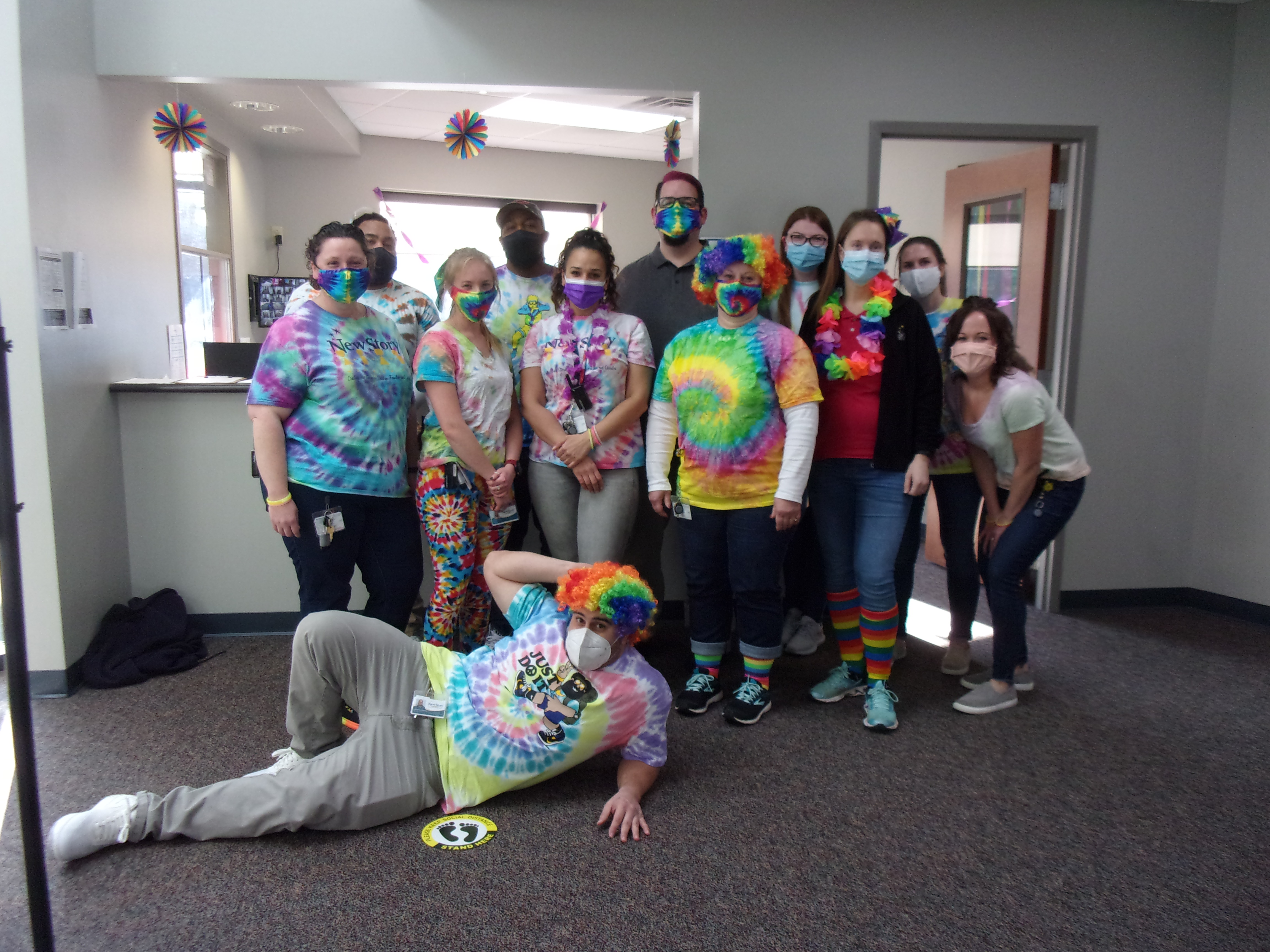 Staff dressed in tie dye, one staff member with a rainbow clown wig on is posed on the floor, the others crouch and stand by in a posed grouping.