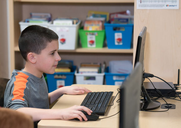 A young boy in a grey and orange shirt works on the computer in his autism support classroom