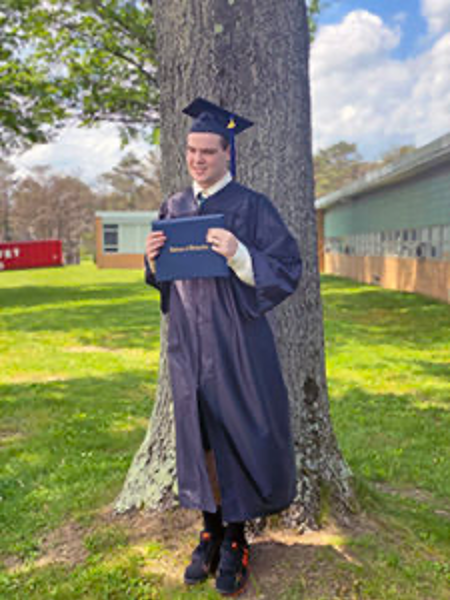 Male graduate in blue cap and gown poses against a tree.