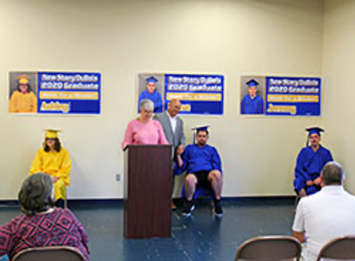 Graduation Ceremony with speaker at podium. Graduates sit on chairs distanced from each other behind podium.